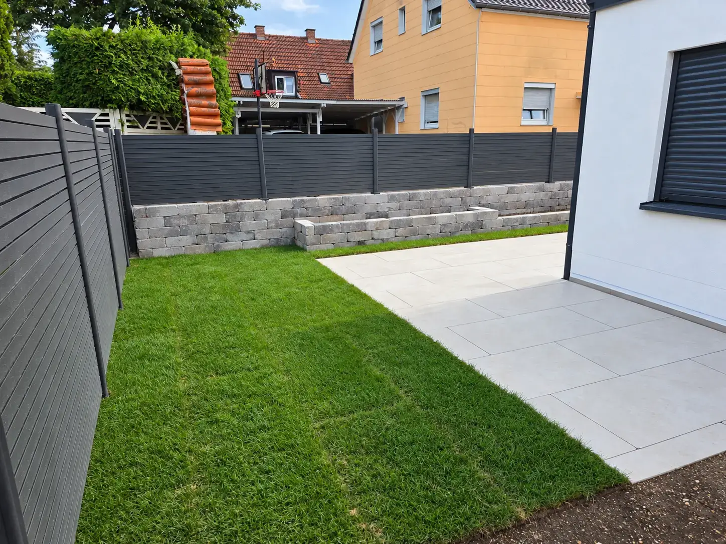 Neu angelegte Rasenfläche und helle Plattenterrasse mit Pflanzmauern und anthrazitfarbenem Sichtschutz