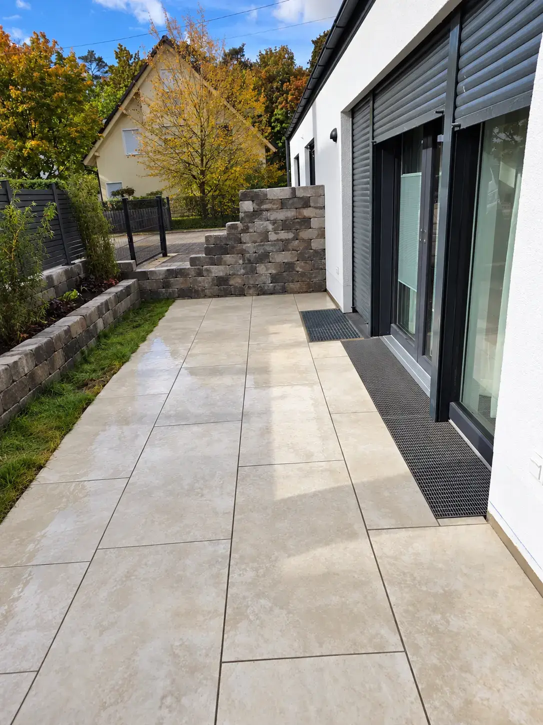 Fertige Terrasse aus hellen großformatigen Platten entlang des Hauses, mit Mauerbeeten und Rasenstreifen